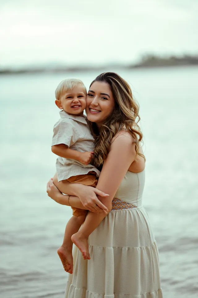 Mãe com o filho pequeno no colo, os dois sorrindo, o mar atrás