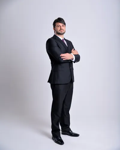 Executivo de terno preto e gravata, braços cruzados, retrato de corpo inteiro em fundo claro
