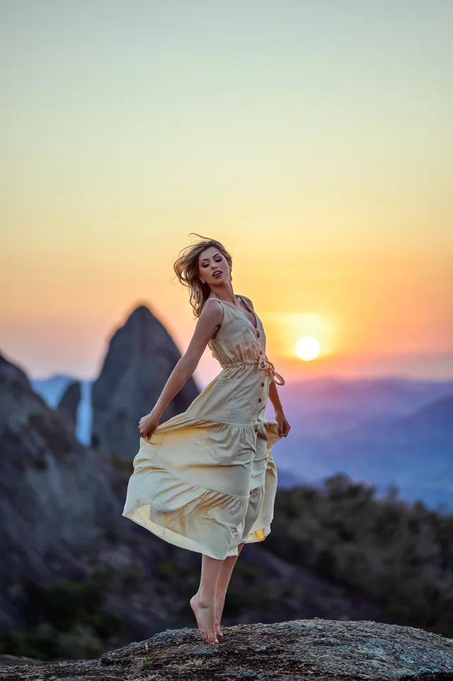 Mulher de vestido claro no alto de uma montanha, contra o sol no fim da tarde