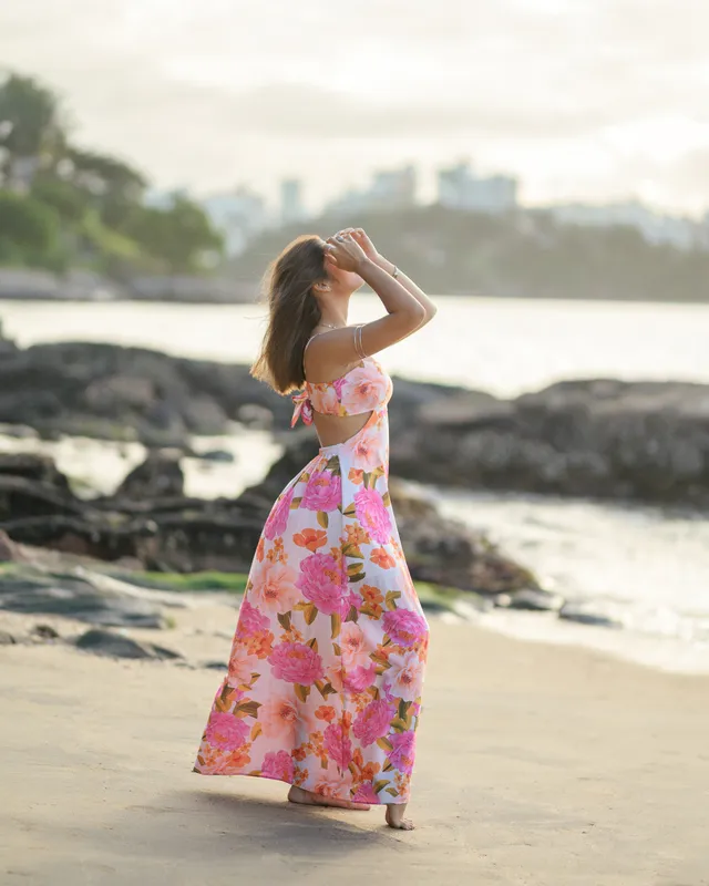 Mulher de vestido florido caminhando na praia, com a cidade ao fundo