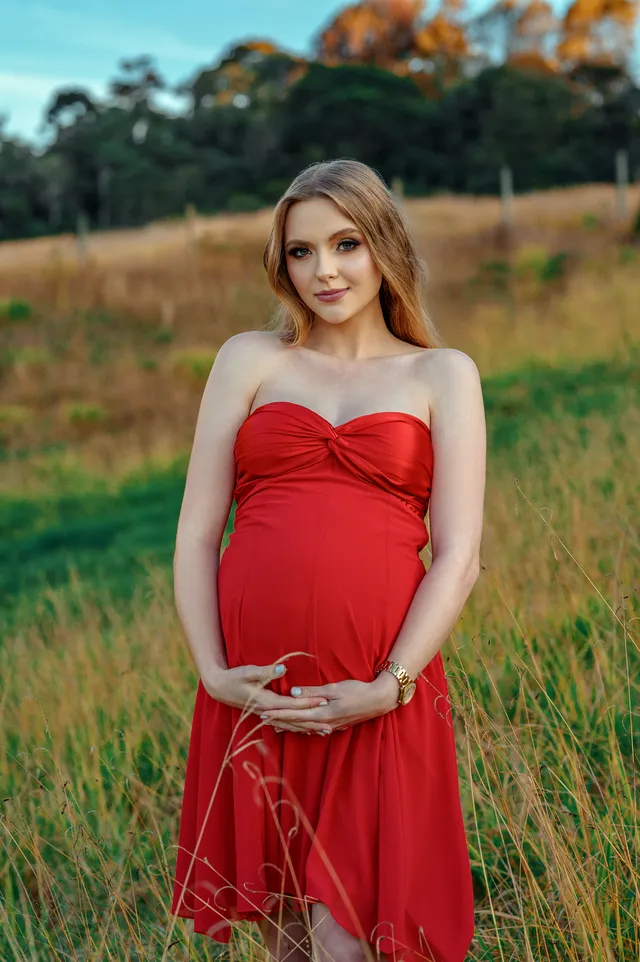 Gestante de vestido vermelho longo em campo aberto, com as mãos na barriga