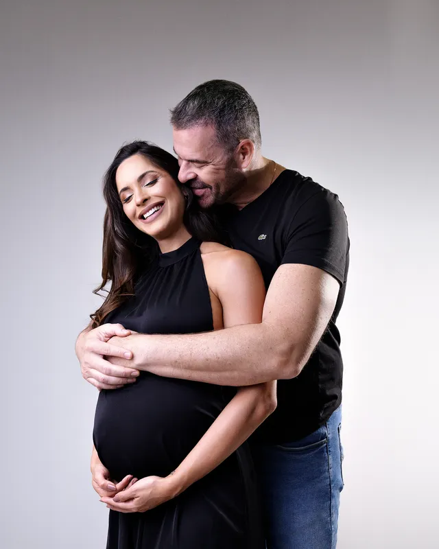 Casal abraçado em estúdio de fundo neutro, as mãos dele sobre a barriga dela